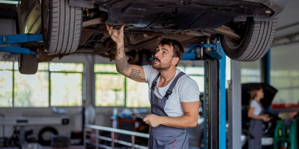 Mechanic holding a wrench while inspecting the underside of a car raised on a hydraulic lift in an auto repair shop
